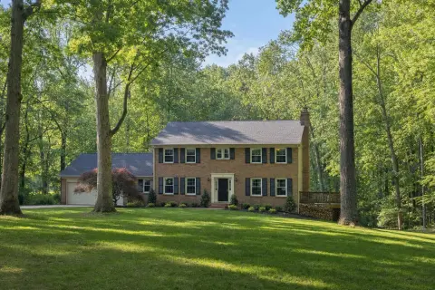 Brick colonial home on a large wooded lot in Derwood Maryland with a back deck, a stacked woodpile, and tall oaks at the edge of the forest