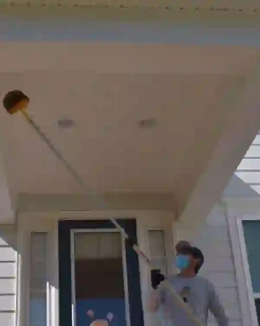 Pest control technician using a webster tool to remove spider webs from porch eaves