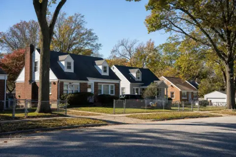 Row of post-war brick and vinyl-sided Cape Cod homes with dormer windows and chain-link fences under autumn oak trees on a residential street in Glen Burnie, Maryland