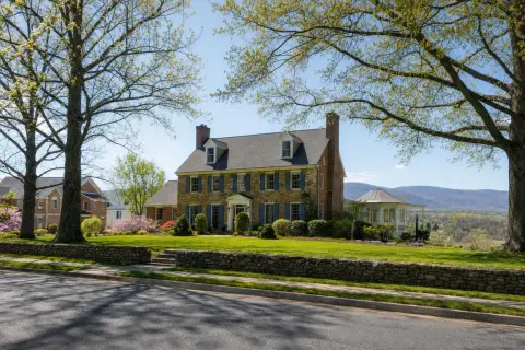 Residential street in Loudoun County Virginia with brick colonial homes, mature oak trees, a stacked fieldstone wall, and the Blue Ridge foothills in the distance