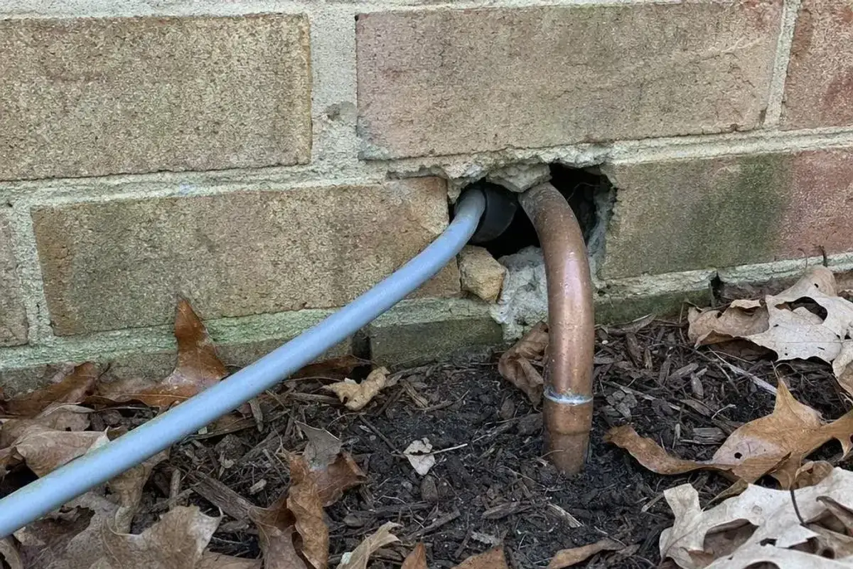 Unsealed gap around a utility cable and copper pipe entering a brick wall near the ground