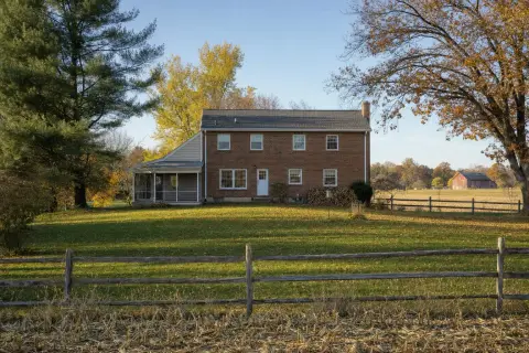 Back lawn of a brick colonial home in Olney, Maryland bordered by a split-rail fence, a harvested cornfield and a red barn in autumn