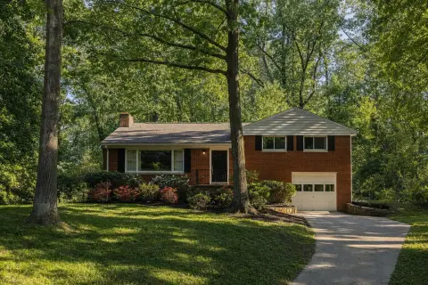 Late-1950s red brick split-level house on a sloping wooded lot under tall oak trees in Silver Spring Maryland