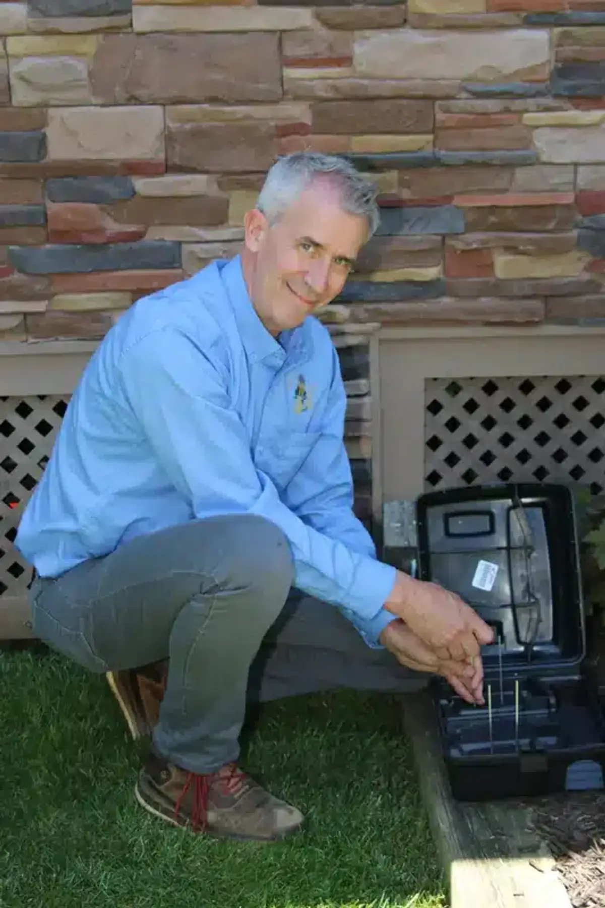 Pest control technician kneeling next to a monitoring station at the foundation of a residence