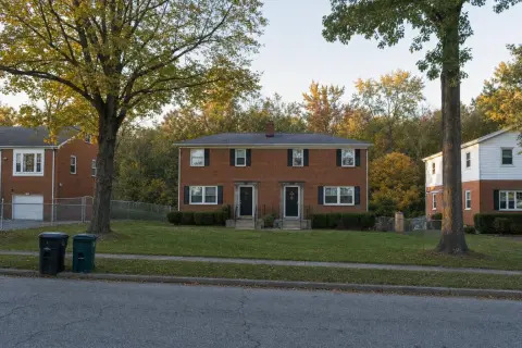 1950s red-brick duplex with two front doors, a shared lawn, and autumn trees on a quiet street in Temple Hills, Maryland